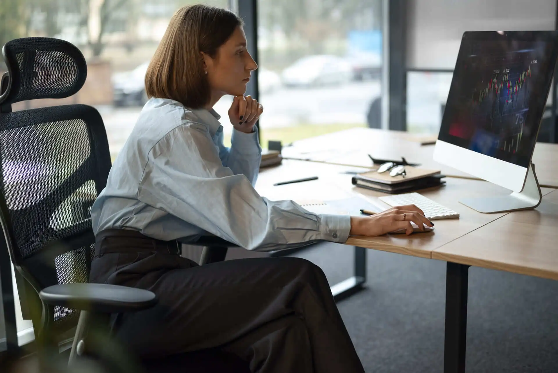 Eine Frau in Business-Kleidung sitzt konzentriert an einem modernen Schreibtisch in einem hellen ergonomischen Büro. Mit einer Hand stützt sie ihr Kinn, während sie mit der anderen die Computermaus bedient. Auf dem Schreibtisch liegen Notizbücher, Stifte und eine Brille. Im Hintergrund sind große Fenster mit Blick auf eine Straße und parkende Autos zu sehen. Die Szene vermittelt eine professionelle und analytische Arbeitsatmosphäre, die sich auf Finanzen oder Datenanalyse konzentriert.