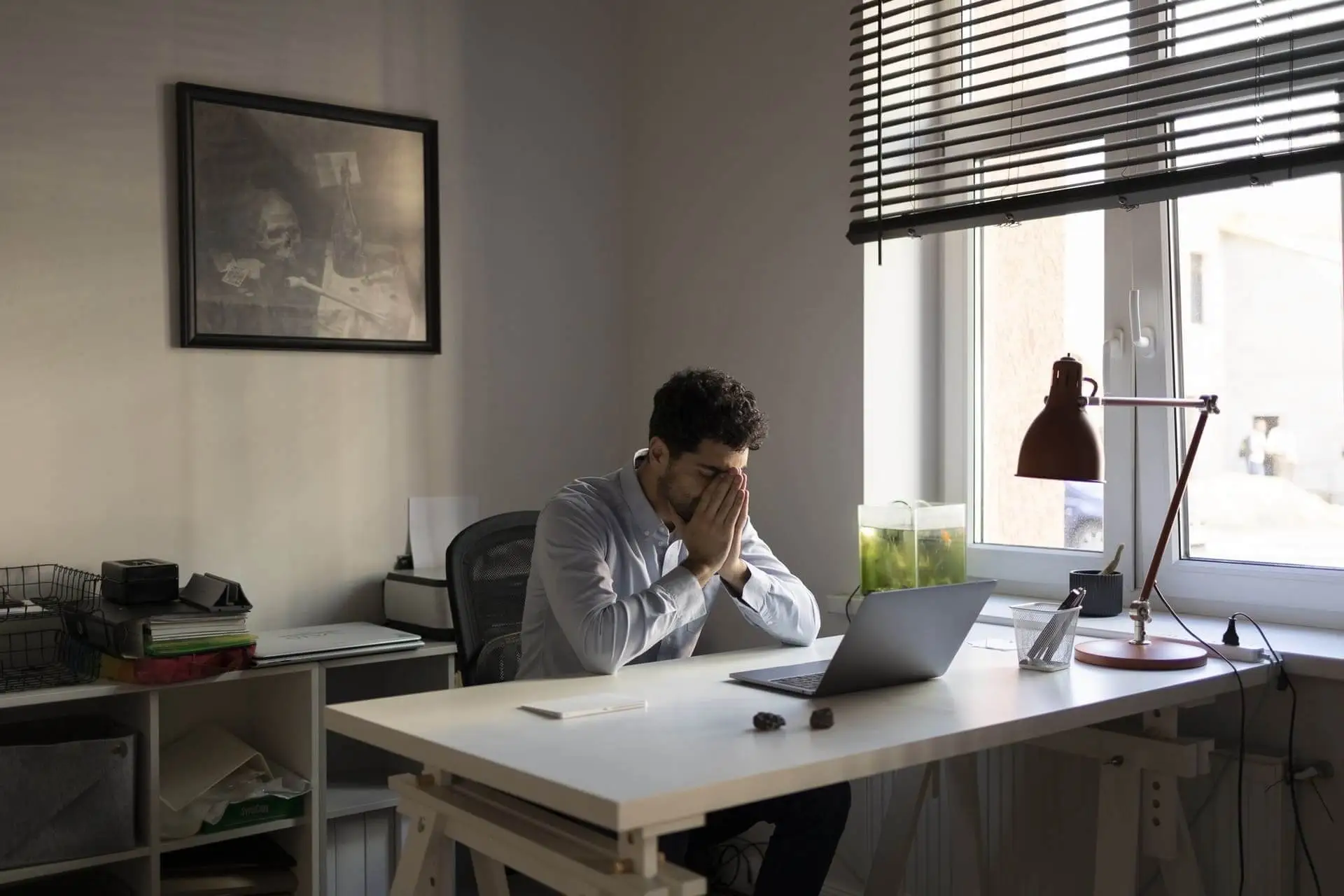 Ein junger Mann sitzt an einem weißen Schreibtisch in einem minimalistischen Büro. Er trägt ein helles Hemd und hält sein Gesicht in den Händen, als wäre er gestresst oder nachdenklich. Vor ihm steht ein aufgeklappter Laptop, daneben liegen ein Smartphone und einige kleine Gegenstände. Die Szene wird durch natürliches Licht von einem großen Fenster erhellt, neben dem eine Schreibtischlampe und ein kleines Aquarium stehen. An der Wand hängt ein düsteres Gemälde mit einem Totenkopf und Symbolen der Vergänglichkeit, was die melancholische Stimmung des Bildes verstärkt.