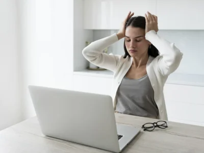 Eine junge Frau mit dunklen Haaren sitzt an einem Holztisch vor einem offenen Laptop und wirkt gestresst. Sie hält ihre Hände an den Kopf und hat einen erschöpften Gesichtsausdruck, was auf psychische Belastung hindeutet. Neben dem Laptop liegt eine schwarze Brille. Der Hintergrund zeigt eine helle, moderne Küche.