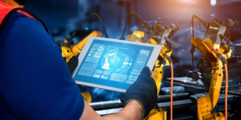 Ein Techniker nutzt ein Tablet, um mit Hilfe von KI in der Produktion die Leistung von Roboterarmen in einer automatisierten Fertigungsanlage zu steuern.