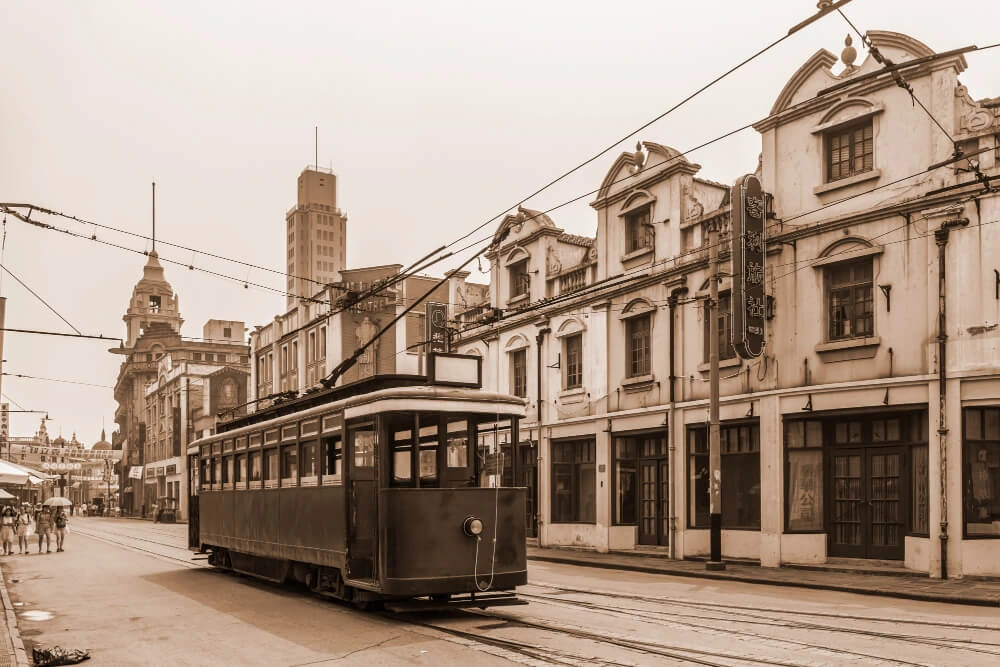 Ein historisches Foto zeigt eine Straßenbahnszene in Shanghai zur Zeit der Industrie 2.0. Eine dunkle, elektrische Straßenbahn fährt auf Schienen durch eine Straße mit mehrstöckigen Gebäuden im Kolonialstil. Stromleitungen überspannen die Straße. Im Hintergrund sind weitere historische Gebäude zu sehen, einschließlich eines Turms.