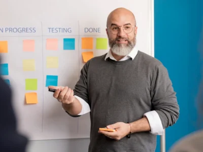 Ein lächelnder Mann mit Brille und grauem Pullover erklärt agile Produktentwicklung anhand eines Taskboards. Auf dem Whiteboard hinter ihm sind die Spalten "To Do", "In Progress", "Testing" und "Done" mit bunten Haftnotizen bestückt. Er hält einen Marker und Haftnotizen in der Hand und spricht zu einem Team, das außerhalb des Bildes sichtbar ist.