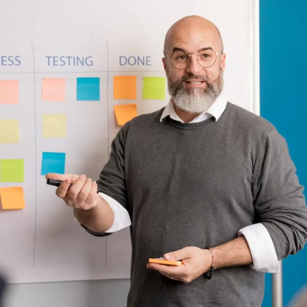 Ein lächelnder Mann mit Brille und grauem Pullover erklärt agile Produktentwicklung anhand eines Taskboards. Auf dem Whiteboard hinter ihm sind die Spalten "To Do", "In Progress", "Testing" und "Done" mit bunten Haftnotizen bestückt. Er hält einen Marker und Haftnotizen in der Hand und spricht zu einem Team, das außerhalb des Bildes sichtbar ist.
