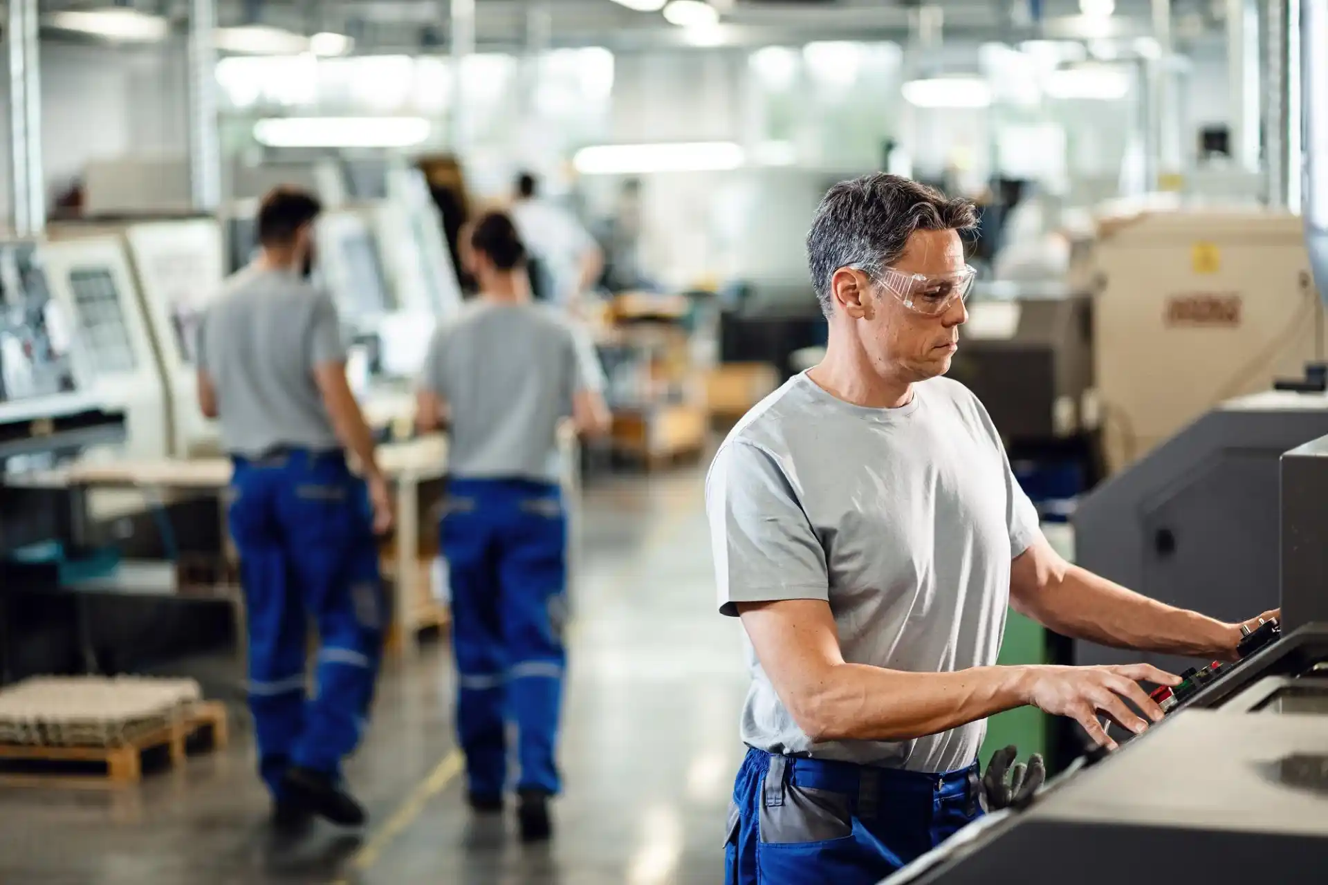 Industriearbeiter bedient eine CNC-Maschine in einer modernen Produktionshalle, während Kollegen im Hintergrund an weiteren Maschinen arbeiten. Alle tragen Schutzbrillen und Arbeitskleidung.