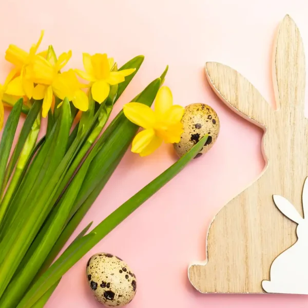 Eine frühlingshafte Szene mit gelben Narzissen, braun gesprenkelten Wachteleiern und einer hölzernen Hasenfigur auf einem rosa Hintergrund. Während dieses Bild die typische Osterdekoration und -feierlichkeiten zeigt, steht es im Kontrast zum Thema Ostermontag arbeiten, da es die Ruhe und Festlichkeit des Feiertags symbolisiert.