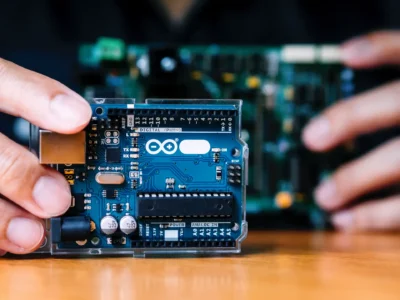 Nahaufnahme eines Arduino-Boards, das als kompakte PLC (Programmierbare Logiksteuerung) in der Hand gehalten wird – im Hintergrund eine weitere Leiterplatte; genutzt für Automatisierungs- und Steuerungsprojekte.