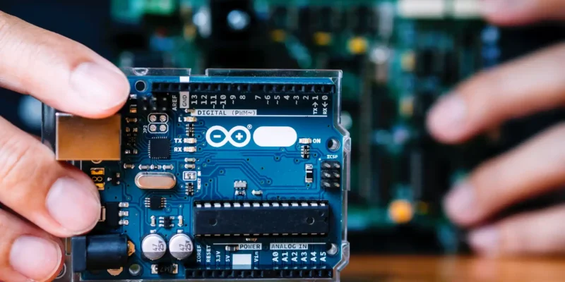 Nahaufnahme eines Arduino-Boards, das als kompakte PLC (Programmierbare Logiksteuerung) in der Hand gehalten wird – im Hintergrund eine weitere Leiterplatte; genutzt für Automatisierungs- und Steuerungsprojekte.