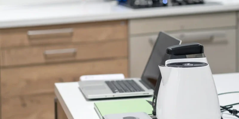 Ein Wasserkocher, ein Laptop und weitere elektronische Geräte stehen auf einem weißen Küchentresen in einer modernen Büroküche. Im Hintergrund sind ein Gasherd, ein Topf und Küchenschränke zu sehen.