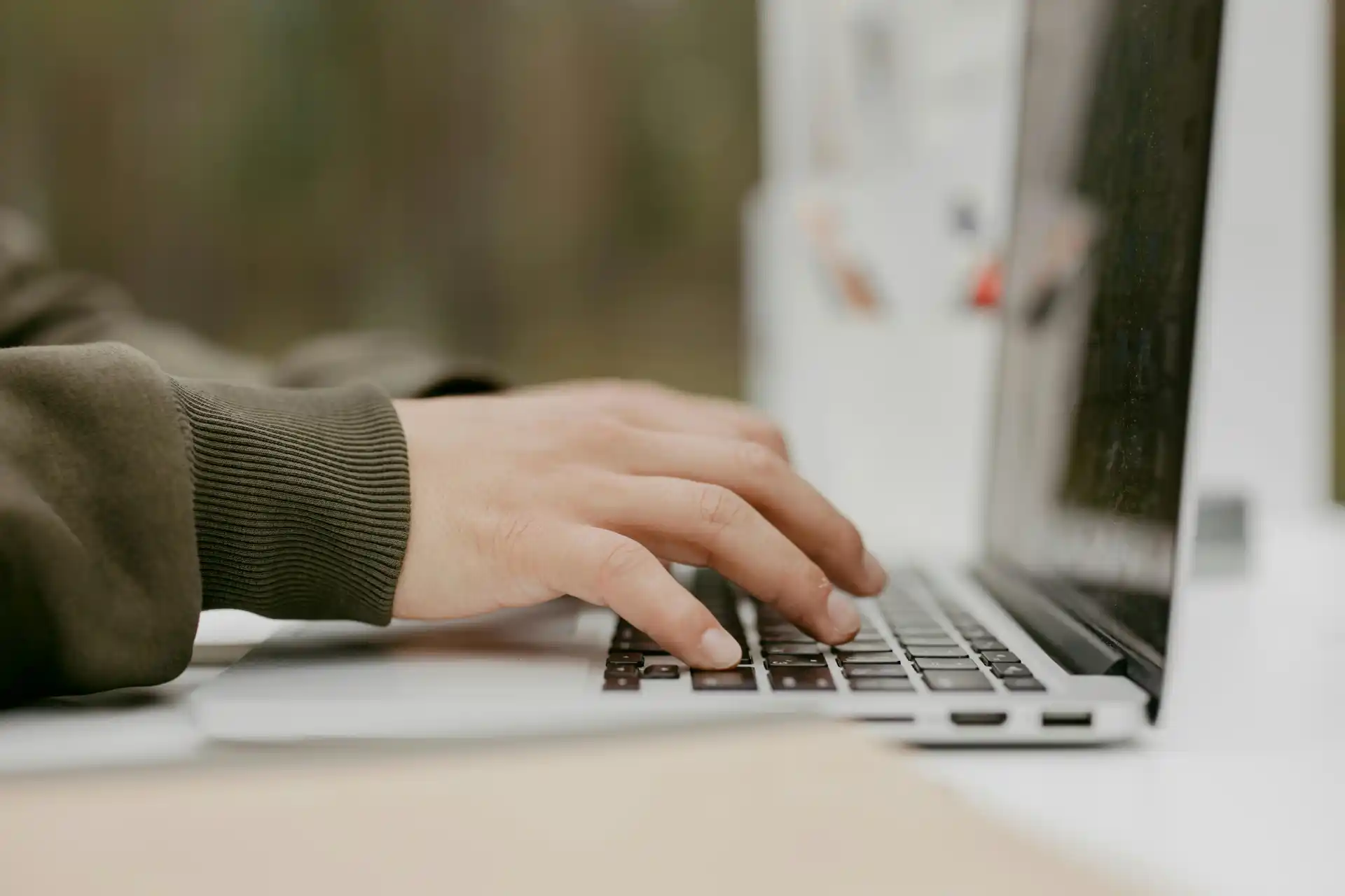 Eine Person tippt auf einer Laptop-Tastatur. Der Fokus liegt auf der rechten Hand, die mittig über der Tastatur schwebt. Die Person trägt einen olivgrünen Pullover. Der Laptop steht auf einem weißen Tisch, im Hintergrund ist ein unscharfer, natürlicher Außenbereich zu erkennen.