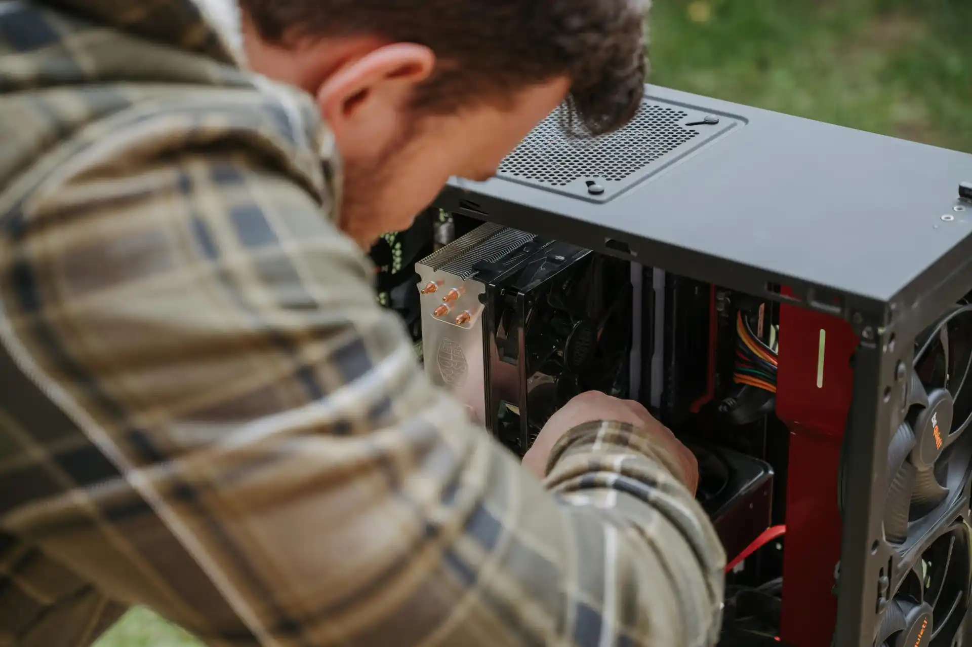 Ein Mann in kariertem Hemd überprüft im Freien die Komponenten eines geöffneten PC-Gehäuses. Das Gerät wird aktuell geprüft, einschließlich CPU-Kühler, RAM und Kabelverbindungen.