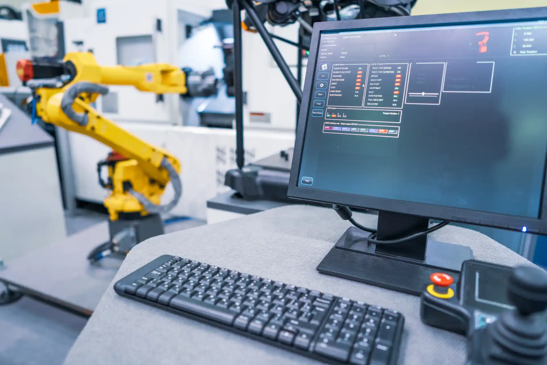 Monitor mit Benutzeroberfläche zur Steuerung eines gelben Industrieroboters in einer automatisierten Produktionszelle – moderne Fertigungstechnologie mit Tastatur, Not-Aus-Schalter und Roboterarm im Hintergrund.