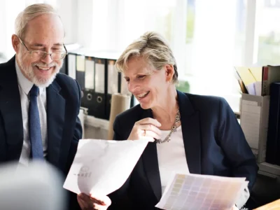 Zwei erfahrene Geschäftsleute im Büro lachen gemeinsam beim Durchsehen von Dokumenten – symbolisiert positive Teamarbeit und den verantwortungsvollen Umgang mit Arbeitszeiten im Rahmen des Arbeitszeitgesetzes.