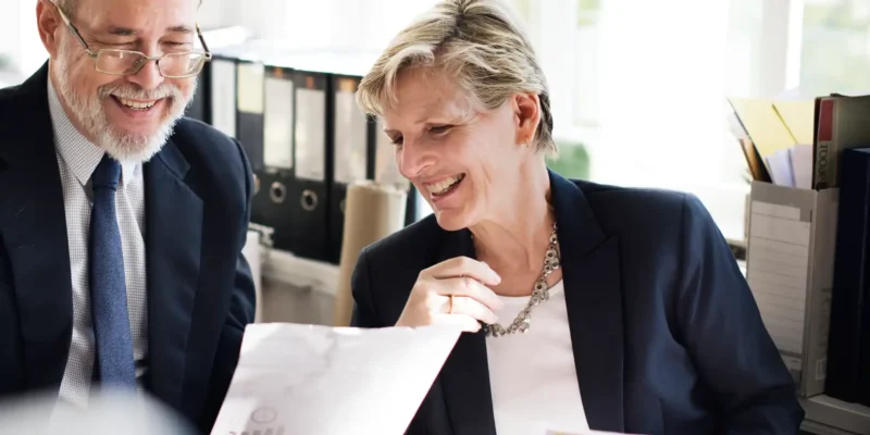 Zwei erfahrene Geschäftsleute im Büro lachen gemeinsam beim Durchsehen von Dokumenten – symbolisiert positive Teamarbeit und den verantwortungsvollen Umgang mit Arbeitszeiten im Rahmen des Arbeitszeitgesetzes.
