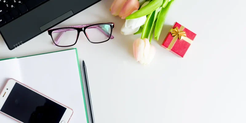 Arbeitsplatz mit Laptop, Smartphone, Brille, Notizbuch und Stift, daneben ein Blumenstrauß und ein kleines Geschenk – Kombination aus Arbeit und festlichem Anlass.