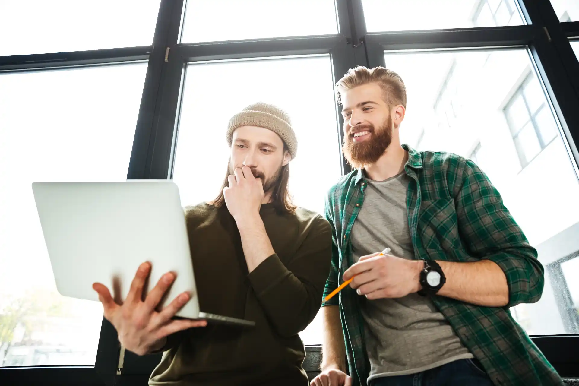 Zwei junge Männer arbeiten konzentriert gemeinsam an einem Laptop in einem hellen Büro – einer hält das Gerät, der andere lächelt und hält einen Stift in der Hand.