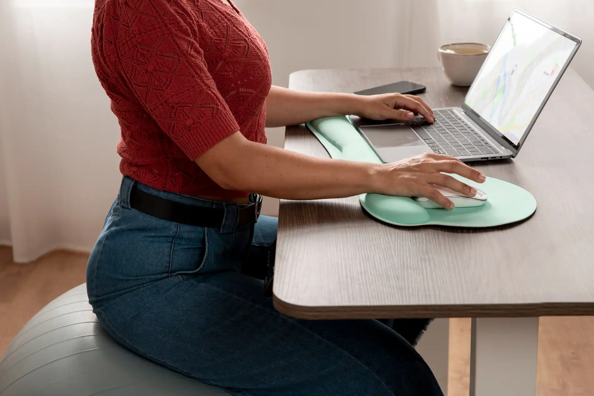 Ein detailliertes Seitenprofil zeigt eine Frau, die an einem modernen Schreibtisch sitzt und konzentriert an einem Laptop arbeitet, was ein Beispiel für ergonomische Arbeitsplatzgestaltung darstellt. Sie sitzt auf einem großen grauen Gymnastikball, der eine dynamische Sitzhaltung fördert. Ihre Hände ruhen auf einer mintgrünen ergonomischen Handballenauflage vor einer passenden Maus und Tastatur, während sie tippt. Sie trägt ein rotes, texturiertes Oberteil und Jeans. Auf dem Schreibtisch sind zudem ein Smartphone und eine Tasse zu sehen. Die Szene vermittelt ein Gefühl von Komfort und bewusster Körperhaltung beim Arbeiten.