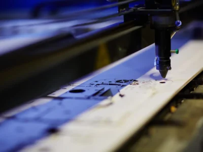 Ein Close-up zeigt den Schneidkopf eines Laser Cutters, der konzentriert und präzise die Oberfläche einer hellen Holzplatte bearbeitet. Der Laserstrahl, sichtbar als roter Punkt, brennt gerade eine feine Linie in das Material. Im Fokus liegen die bereits fertigen und teils herausgeschnittenen Teile links, die die Präzision und Effizienz des Werkzeugs unterstreichen, während der Rest der Maschine im unscharfen Hintergrund verschwindet.