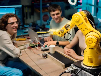 Ein gelber, kleiner Gelenkarmroboter (Cobot) in einem Lab- oder Werkstatt-Setting ist auf einem Tisch montiert und hält einen kleinen, schwarzen Greifer. Drei junge Erwachsene sitzen um den Tisch herum und beobachten den Roboter. Einer der Männer trägt eine Brille und blickt lächelnd auf die Szene, während der andere Mann auf einem Laptop arbeitet, der mit der Robotersteuerung verbunden ist. Dieses Bild visualisiert ein Studien- oder Entwicklungsprojekt im Bereich der Mensch-Roboter-Kollaboration (MRK Abkürzung), bei dem Mensch und Maschine im selben Arbeitsbereich agieren.