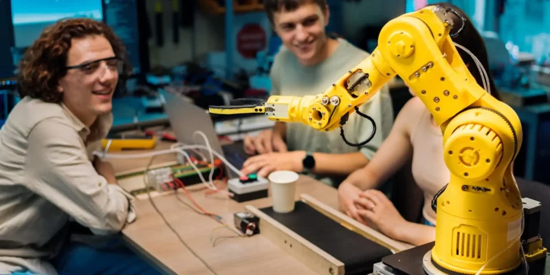 Ein gelber, kleiner Gelenkarmroboter (Cobot) in einem Lab- oder Werkstatt-Setting ist auf einem Tisch montiert und hält einen kleinen, schwarzen Greifer. Drei junge Erwachsene sitzen um den Tisch herum und beobachten den Roboter. Einer der Männer trägt eine Brille und blickt lächelnd auf die Szene, während der andere Mann auf einem Laptop arbeitet, der mit der Robotersteuerung verbunden ist. Dieses Bild visualisiert ein Studien- oder Entwicklungsprojekt im Bereich der Mensch-Roboter-Kollaboration (MRK Abkürzung), bei dem Mensch und Maschine im selben Arbeitsbereich agieren.
