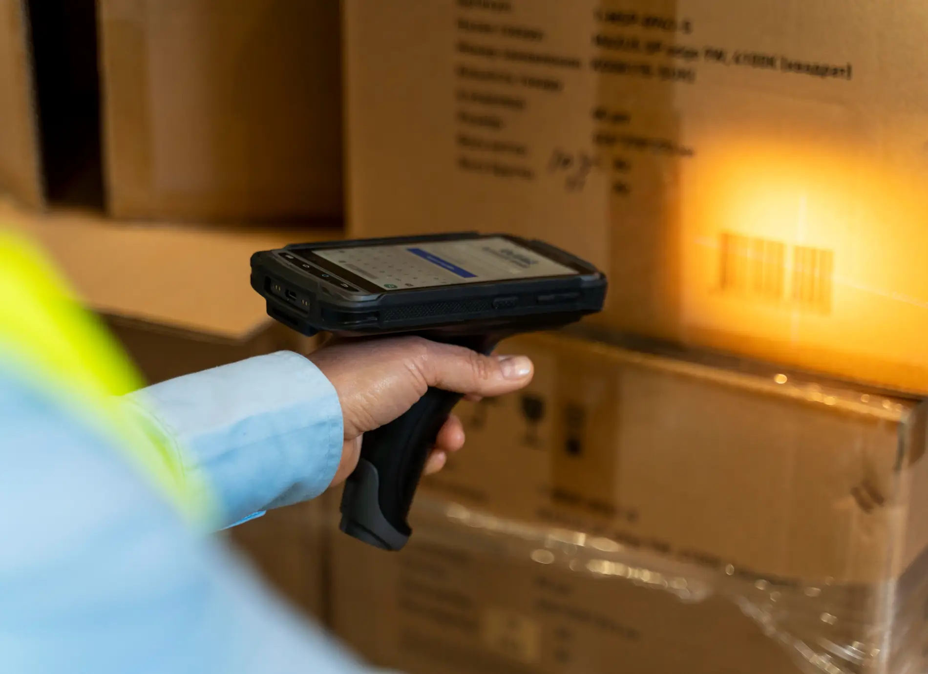 Ein Mitarbeiter im Lager hält ein industrielles Hand-Terminal (Barcode-Scanner) und scannt den Barcode auf einem braunen Versandkarton. Die Szene ist unscharf im Hintergrund, wobei der leuchtende Scan-Balken über dem Code sichtbar ist. Die Hand des Mitarbeiters mit einem Teil des Ärmels einer hellblauen Uniform und einer gelben Warnweste ist im Vordergrund zu sehen. Dieses Bild symbolisiert die Datenerfassung in der Logistik, die ein wichtiger, aber manueller Schritt ist, der durch ein automatisiertes Pick and Place System abgelöst oder ergänzt werden könnte, um die Effizienz der Warenbewegung zu erhöhen.